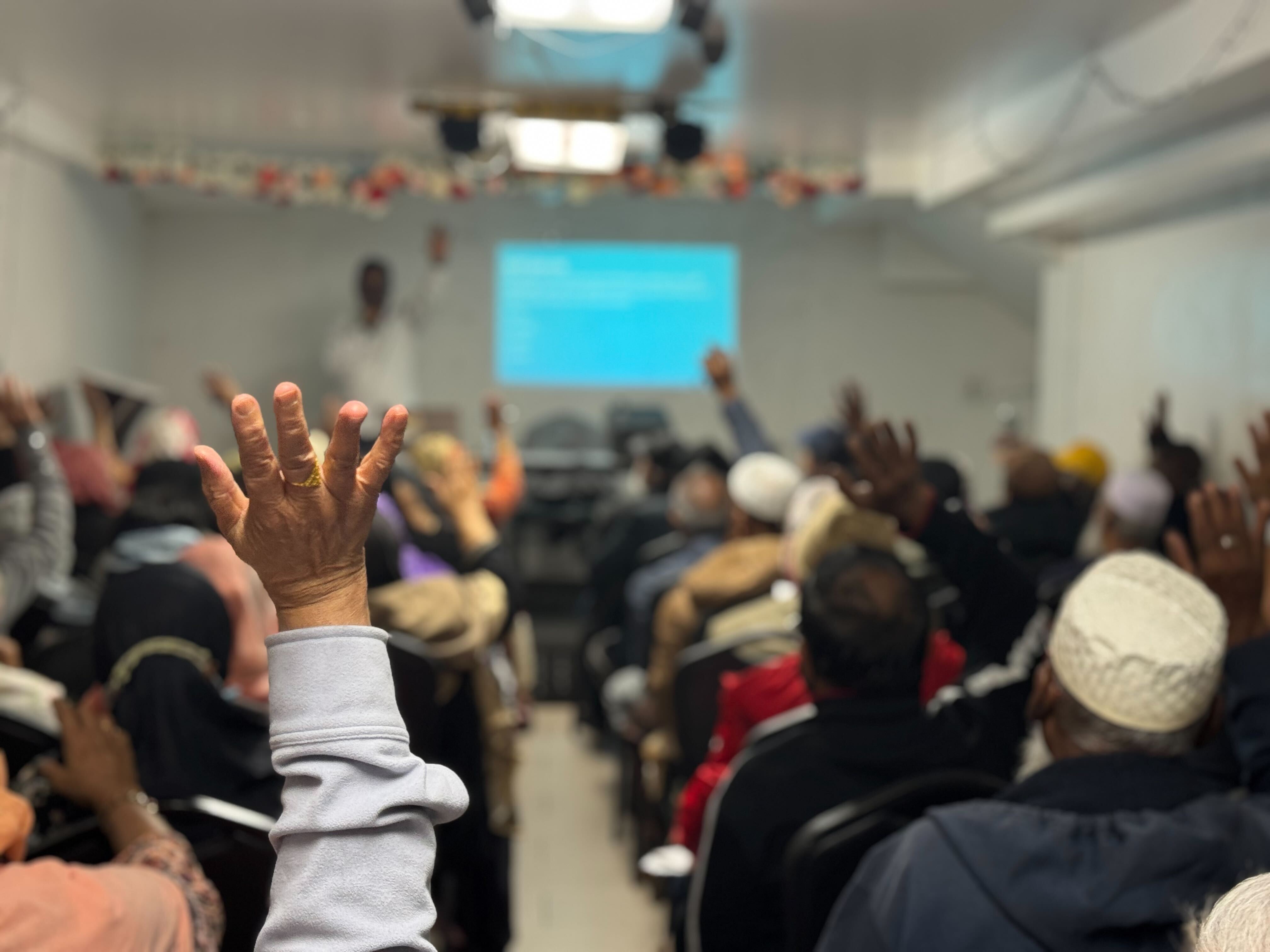 A room of people seated viewing a presentation with a close up on a raised hand 
