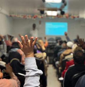 A room of people seated viewing a presentation with a close up on a raised hand 