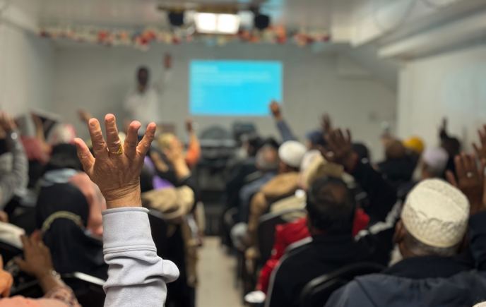 A room of people seated viewing a presentation with a close up on a raised hand 